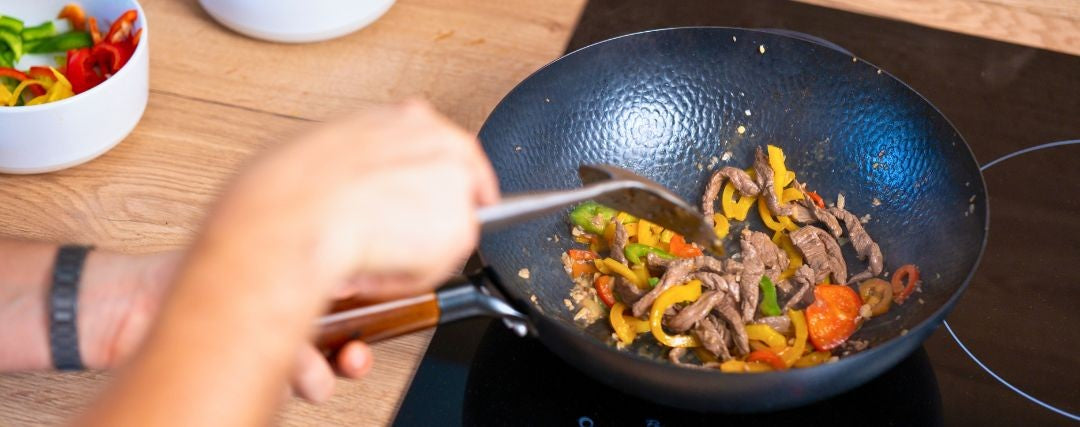 Hand stirring beef strips and sliced peppers in a hammered wok on an induction hob, next to a bowl of peppers Image